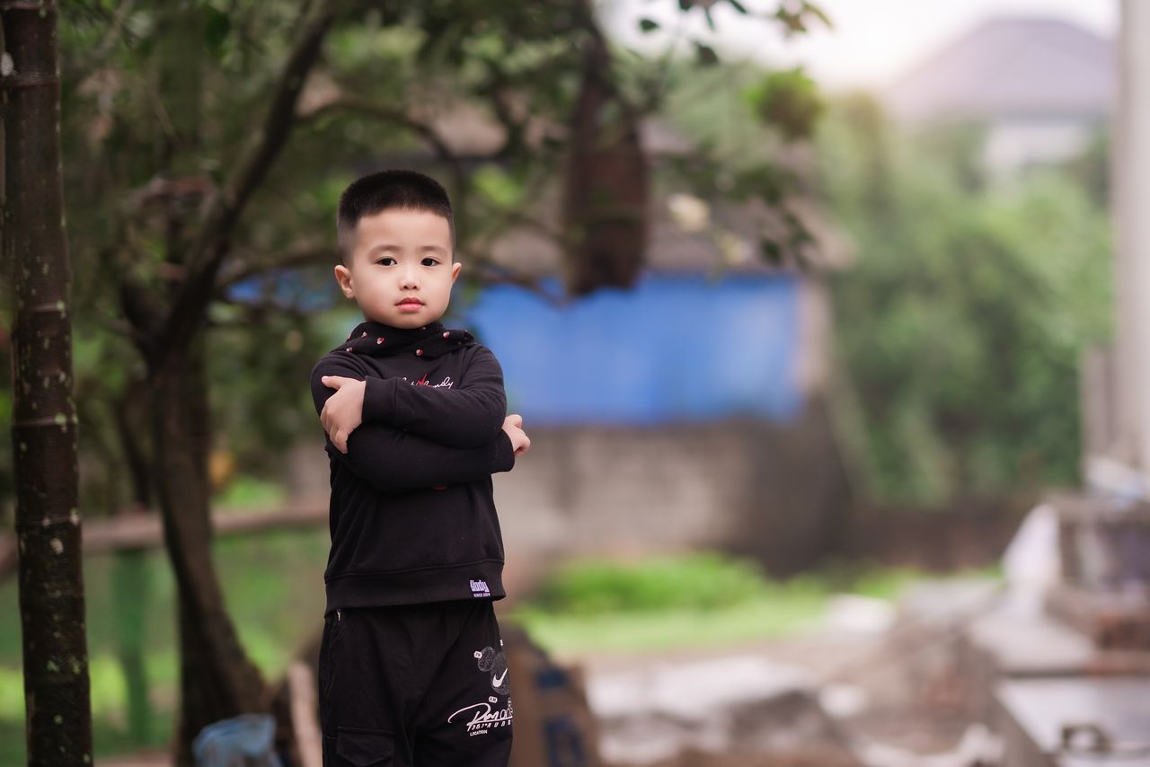 Services A confident young boy standing outdoors, arms crossed, wearing trendy fashion, in a natural setting.
