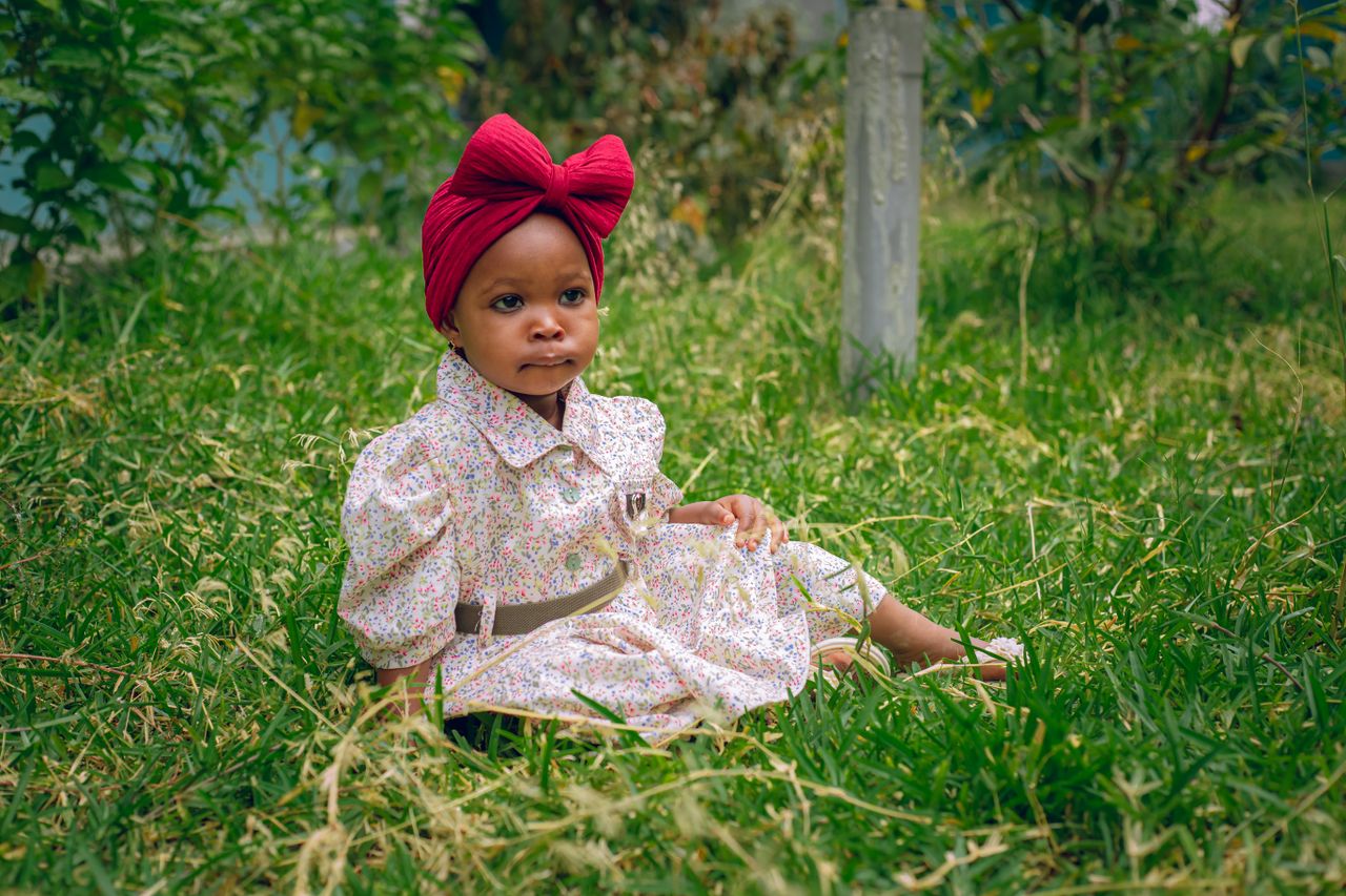 Services Adorable toddler sitting in a garden wearing a floral dress and red headband, depicting innocence and joy.