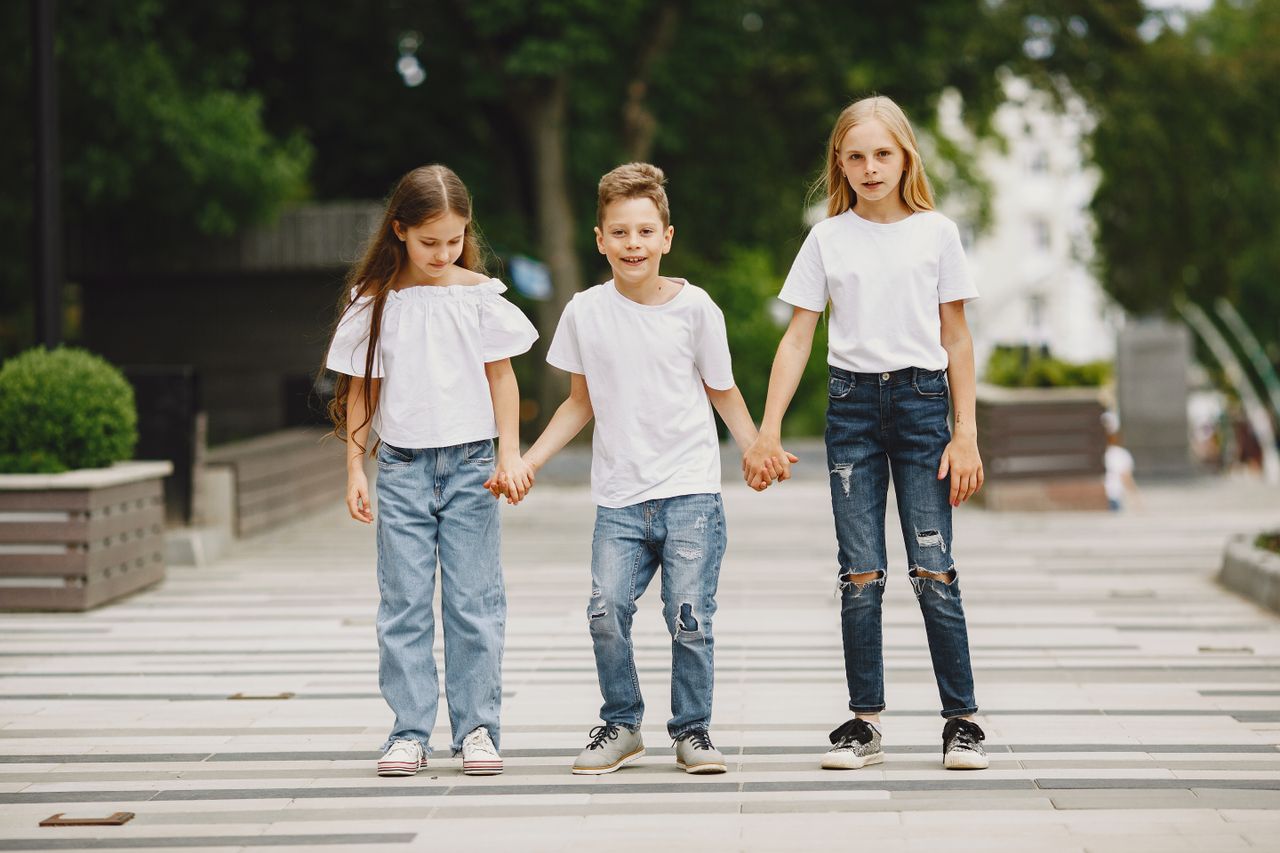 Three children in white t-shirts and jeans holding hands outdoors on a summer day.