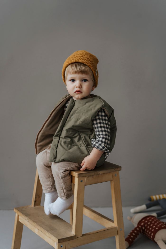 Services Cute child wearing knit cap and vest, sitting on a wooden chair in a studio setting.