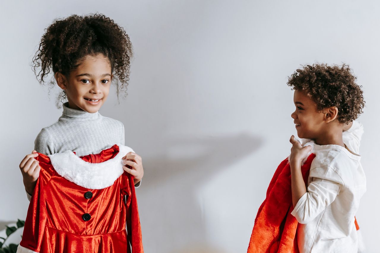 Services Cheerful African American siblings with red Santa Claus outfits in hands standing in light room at home during Christmas holiday